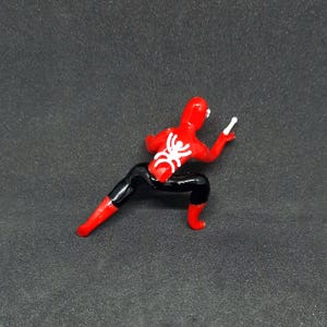 May include: A red and black Spider-Man action figure in a crouching pose. The figure has a white spider emblem on its back and is holding a white object in its right hand. The figure is on a dark gray surface.