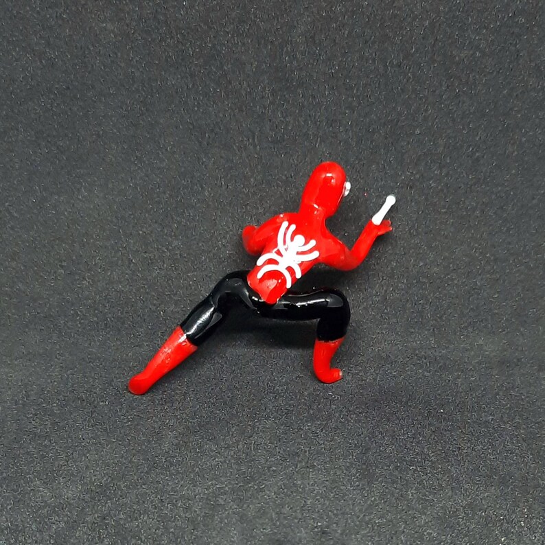 May include: A red and black Spider-Man action figure in a crouching pose. The figure has a white spider emblem on its back and is holding a white object in its right hand. The figure is on a dark gray surface.