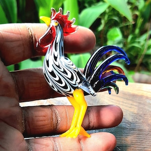 Può includere: Una statuetta di gallo in vetro colorato con vortici neri e bianchi sul corpo, una cresta e bargigli rossi e una coda blu e arancione.