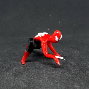 May include: A red and black glass figurine of a person in a crawling position. The figurine has white accents on the head and back. The figure is on a dark surface.