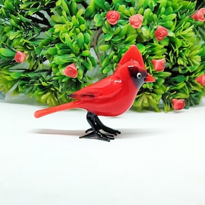 May include: A red cardinal bird figurine made of glass with black eyes and legs. The figurine is perched on a white surface.