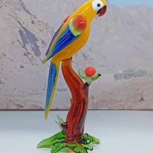 May include: A glass figurine of a yellow, blue and red parrot perched on a brown branch with a red berry. The parrot has a black beak and white eyes.