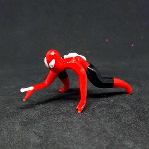 May include: A small, red glass figurine in a crawling pose, with black shorts and white accents on the head and hands. The figurine is on a dark surface.