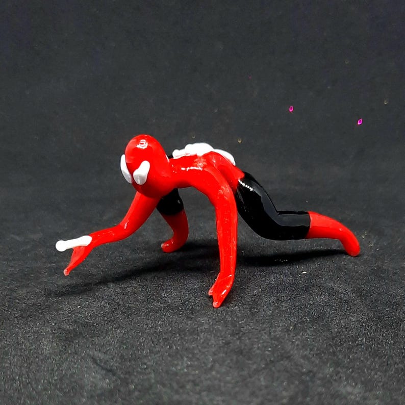 May include: A small, red glass figurine in a crawling pose, with black shorts and white accents on the head and hands. The figurine is on a dark surface.