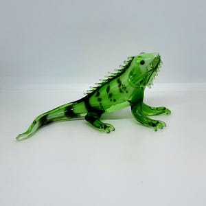 Puede incluir: Una figura de iguana de vidrio verde translúcido con una cresta espinosa a lo largo de la espalda, una cola larga y afilada y ojos pequeños de color negro. El cuerpo presenta manchas moteadas de color verde oscuro y un acabado pulido.
