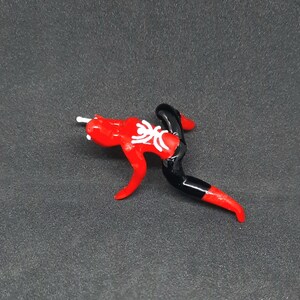 May include: A red and black glass figurine in the shape of a stylized figure with a white floral design on the chest. The figurine has a white rod extending from the head.