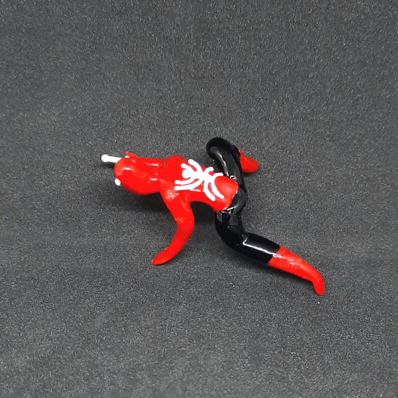 May include: A red and black glass figurine in the shape of a stylized figure with a white floral design on the chest. The figurine has a white rod extending from the head.