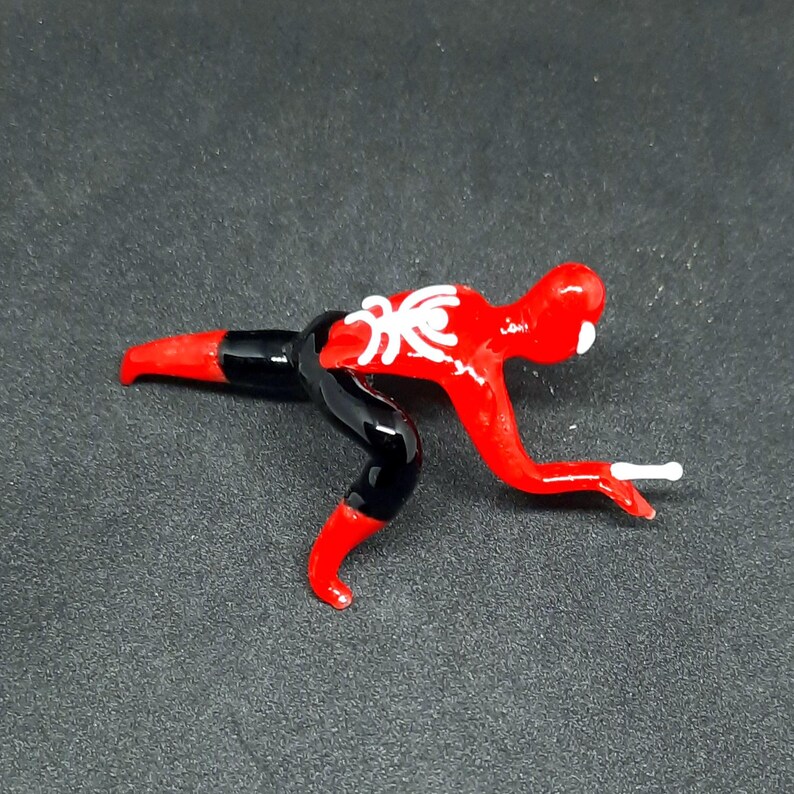 May include: A red and black glass figurine of a person in a crouching position. The figurine has a white spider emblem on the chest and a white detail on the head. The figure is on a dark gray surface.