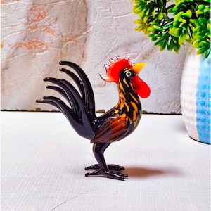 Könnte beinhalten: Eine Glashahnfigur mit schwarzem Körper und bernsteinfarbenen Akzenten an Hals und Flügeln. Der Hahn hat einen leuchtend roten Kamm und Kehllappen, einen gelben Schnabel und einen langen, geschwungenen schwarzen Schwanz aus glattem Glas.