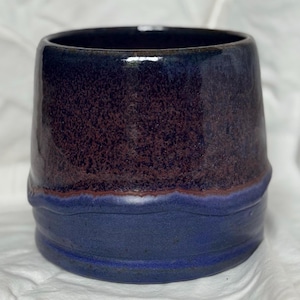 May include: A handmade ceramic mug with a dark blue rim and base, and a textured, speckled brown and purple midsection. The mug has a unique, slightly flared shape and a glossy finish.