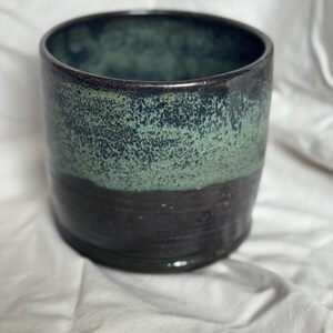 Puede incluir: Una maceta cilíndrica de cerámica con interior verde oscuro y exterior texturizado, moteado en verde y negro. La maceta tiene un borde marrón oscuro y una base negra oscura. La cerámica hecha a mano es adecuada para uso en interiores o exteriores.