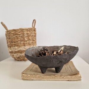 May include: A dark gray, three-legged decorative bowl filled with pine cones and dried botanicals. The bowl sits on a stack of aged paper, with a woven basket in the background. The bowl has a textured surface.