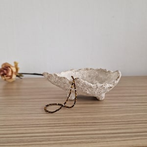 Op de afbeelding: Een getextureerde, gebroken witte decoratieve kom met drie kleine voetjes. Een bruine kralenketting ligt in de kom. Een gedroogde roos ligt op het houten oppervlak naast de kom. De kom wordt waarschijnlijk gebruikt voor sieraden of kleine voorwerpen.