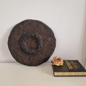 Pode incluir: Um objeto decorativo grande, redondo e castanho escuro com um centro circular em relevo. Uma rosa seca e um livro com letras douradas estão ao lado. O objeto tem uma superfície rugosa e texturizada.