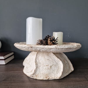 May include: White ceramic pedestal with a scalloped edge, holding two white candles and pine cones.