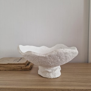 May include: White ceramic bowl with a textured, organic shape and a wide, shallow base. The bowl is on a wooden surface.