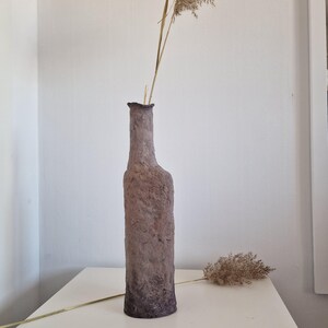Peut inclure: Un vase en forme de bouteille, texturé, de couleur marron et grise. Le vase a un col étroit et une surface rugueuse et inégale. Des herbes séchées sont placées à l'intérieur du vase. Le vase est posé sur une surface blanche.