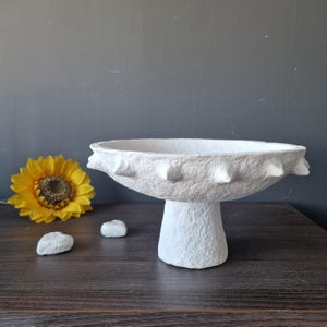 Puede incluir: Un cuenco blanco texturizado sobre pedestal con un borde ancho y festoneado. El cuenco se asienta sobre una base corta y redondeada. Un girasol amarillo y pequeñas piedras blancas están dispuestas cerca.