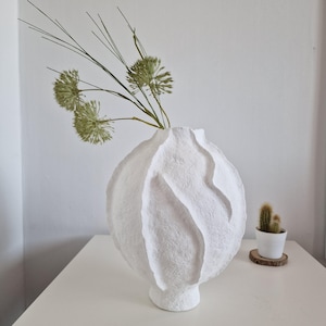 May include: A white, textured vase with a distinctive, rounded form and vertical ridges. It contains green, dried floral stems. A small cactus in a white pot rests on a wooden coaster to the right.