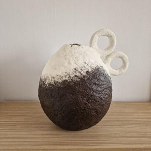 Puede incluir: Un jarrón texturizado hecho a mano con un diseño de dos tonos. La parte inferior es marrón oscuro, con una transición a una sección superior blanca. Dos asas blancas y circulares se extienden desde el lateral. El jarrón está sobre una superficie de madera.