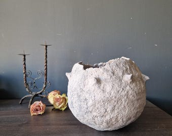 May include: A textured, light gray, spherical decorative bowl with a rough, uneven surface. The bowl has three small, protruding knobs and an irregular opening at the top. A wrought iron candle holder and dried roses are in the background.
