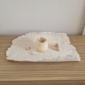 Puede incluir: Una bandeja rectangular blanca y texturizada con un borde irregular. Una vela pequeña usada, dos conchas marinas y un trozo de coral están dispuestos en la bandeja. La bandeja está sobre una superficie de madera.