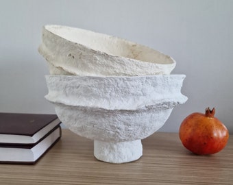 May include: Two white paper mache bowls stacked on top of each other. The bowls have a textured, rough surface and a rounded shape. The bowls are sitting on a wooden table next to a red pomegranate.