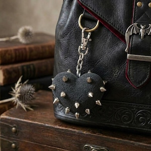 Könnte beinhalten: Ein schwarzer herzförmiger Schlüsselanhänger mit silberfarbenen Metallspitzen an einer schwarzen Ledertasche. Die Tasche verfügt über eine silberfarbene Metallschnalle mit fledermausförmigem Detail und rotem Innenfutter, befestigt mit einem Karabiner.