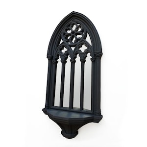 May include: Black gothic-style wall mirror with an arched top and decorative floral and cross accents. The mirror has a small shelf at the bottom and is designed to resemble a cathedral window. The frame is made of a dark material.