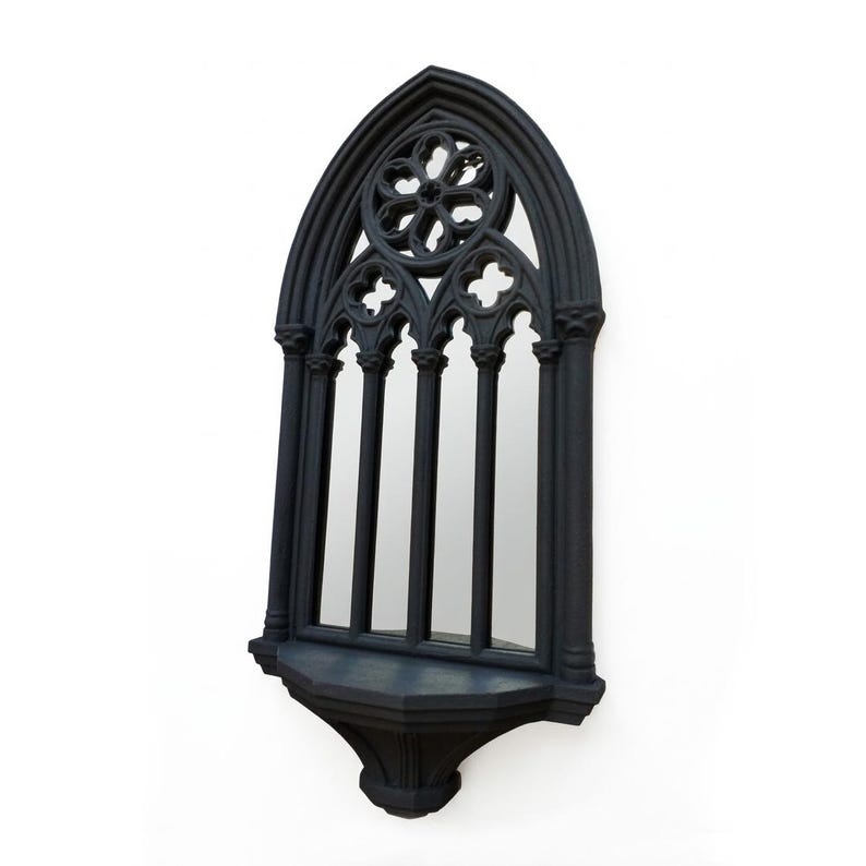 May include: Black gothic-style wall mirror with an arched top and decorative floral and cross accents. The mirror has a small shelf at the bottom and is designed to resemble a cathedral window. The frame is made of a dark material.