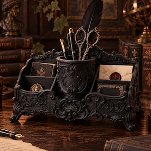 May include: A black desk organizer featuring intricate scrollwork and floral relief patterns. The unit includes a central cylindrical pen holder, side compartments for stationery, and holds a black feather quill, two pens, and ornate metal scissors.