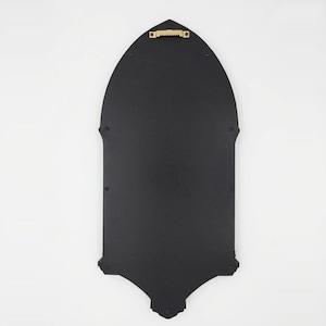 May include: A black, gothic-style wall decor piece with a pointed arch shape. The back features a gold-colored sawtooth hanger for easy mounting. The piece has decorative elements at the top and bottom.