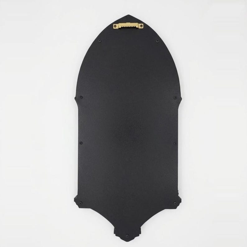 May include: A black, gothic-style wall decor piece with a pointed arch shape. The back features a gold-colored sawtooth hanger for easy mounting. The piece has decorative elements at the top and bottom.