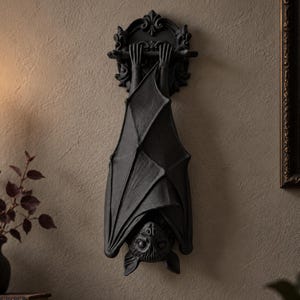 May include: A matte black wall-mounted bat sculpture featuring a bat hanging upside down by its claws from a horizontal bar. The bat has detailed wings folded around its body and a textured face. Above the bar is an ornate, circular decorative mount.