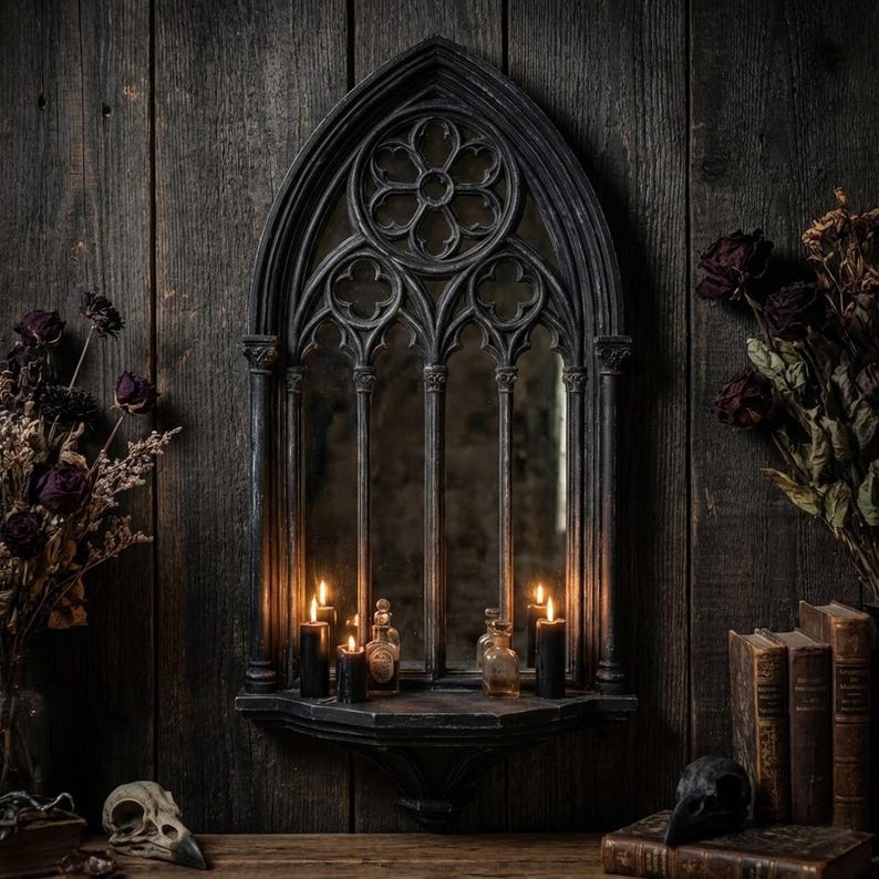 May include: A gothic-style mirror with a dark, ornate frame, featuring a quatrefoil design at the top. The mirror is set against a dark wood background, with lit candles and small bottles on a shelf below. Dried flowers and books add to the vintage aesthetic.