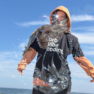 May include: A person in a black t-shirt with the words "brb vibe coding" and an orange hoodie. The image has a surreal effect with a face overlay and water-like textures. The background shows a blue sky and the sea.