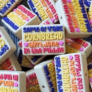 May include: A close-up of several colorful silicone keychains with the phrase "some of y'alls cornbread ain't done in the middle" printed on them. The keychains are made of a soft, flexible silicone material and feature a variety of bright colors, including yellow, pink, blue, and orange.