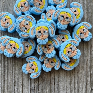 May include: A collection of light blue bunny-shaped charms with yellow hair and sad facial expressions. The charms are made of a rubbery material and are scattered on a wooden surface. The bunny charms have large ears and are wearing a blue hood.