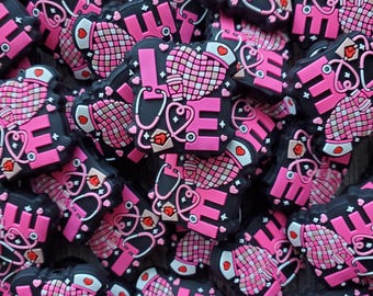 Op de afbeelding: Een close-up van een stapel zwarte en roze siliconen bedels met het woord "LOVE", een stethoscoop en een hartontwerp. De bedels zijn perfect om een ​​persoonlijke touch toe te voegen aan je favoriete accessoires.
