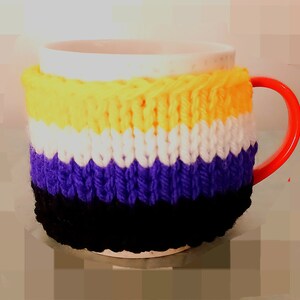 Op de afbeelding: Een witte keramische mok met een rode oor is verpakt in een gebreide hoes. De hoes heeft horizontale strepen in geel, wit, paars en zwart. De mok is ontworpen om dranken warm te houden.