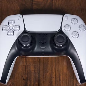 May include: A white and black PlayStation 5 DualSense wireless controller with a black PlayStation logo on the front. The controller has a touchpad, two analogue sticks, and a D-pad.