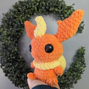 Może przedstawiać: Pluszowa zabawka wykonana na szydełku w kształcie Pok&eacute;mona typu ognistego. Zabawka jest pomarańczowa z ż&oacute;łtymi akcentami i ma duże czarne oczy.