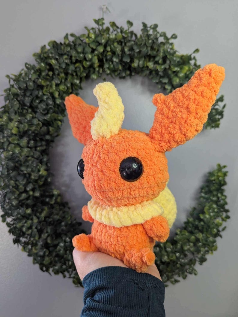 Może przedstawiać: Pluszowa zabawka wykonana na szydełku w kształcie Pok&eacute;mona typu ognistego. Zabawka jest pomarańczowa z ż&oacute;łtymi akcentami i ma duże czarne oczy.