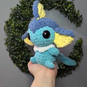 Może przedstawiać: Pluszowa zabawka w kolorze niebieskim i ż&oacute;łtym, wykonana na szydełku, przypominająca wodnego Pokemona. Zabawka ma duże oczy i biały kołnierzyk.