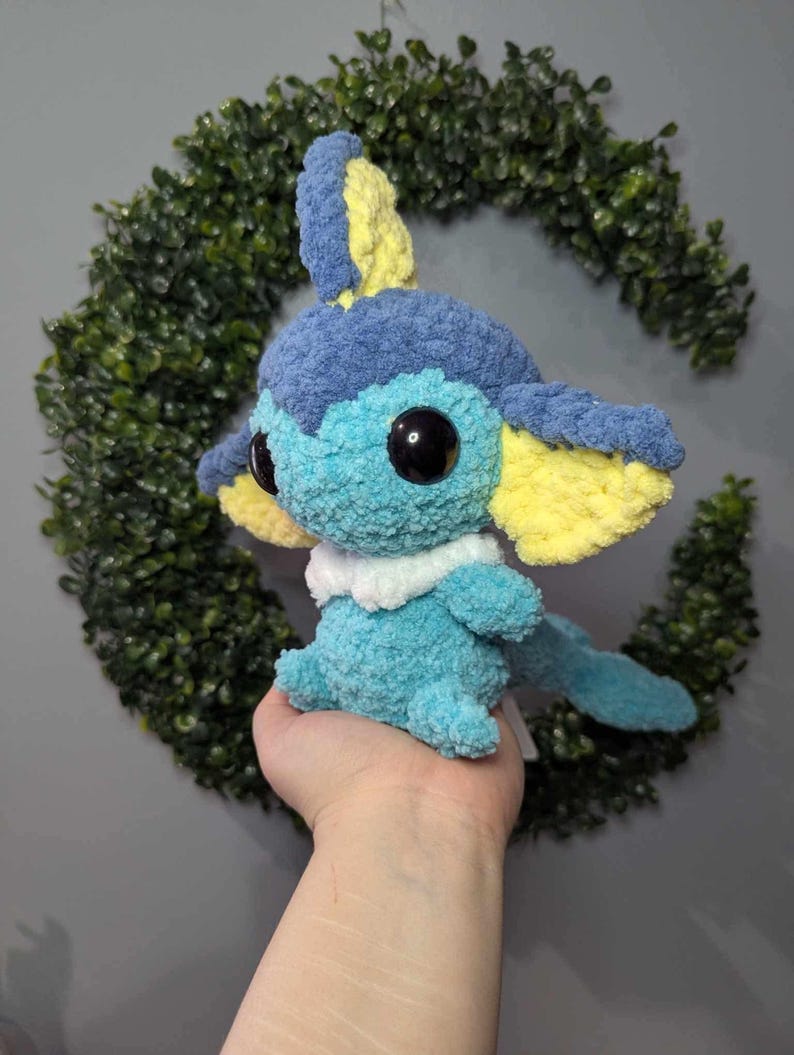 Może przedstawiać: Pluszowa zabawka w kolorze niebieskim i ż&oacute;łtym, wykonana na szydełku, przypominająca wodnego Pokemona. Zabawka ma duże oczy i biały kołnierzyk.