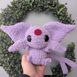 Może przedstawiać: Pluszowa zabawka Eevee wykonana na szydełku w kolorze fioletowym z czerwoną ozdobą na czole i czarnymi oczami. Zabawka jest trzymana w dłoni osoby.