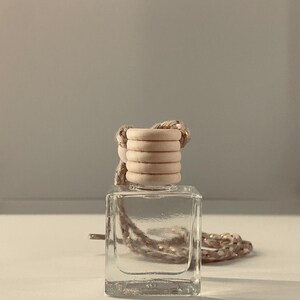 May include: A clear, square glass bottle with a wooden cap and a rope. The cap is made of stacked wooden rings. The rope is a light brown color. The bottle is sitting on a white surface.