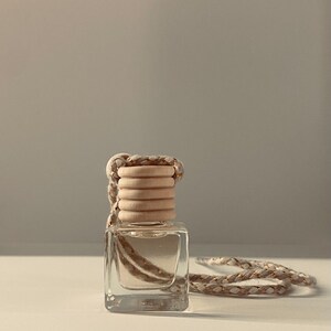 May include: A small, clear glass bottle with a wooden cap and a braided rope. The bottle is filled with a clear liquid. The rope is beige and gold. The product is likely an air freshener or fragrance diffuser.