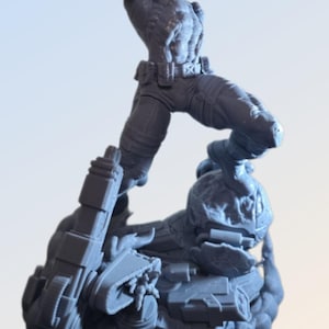 May include: A gray figurine of a muscular figure in a dynamic pose, wearing pants, a belt, and a red head covering. The figure is standing on a detailed base with mechanical elements. The overall design suggests a science fiction or action theme.
