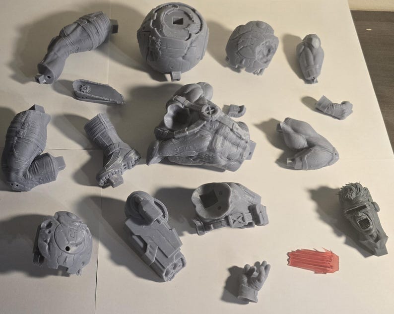 May include: A collection of gray 3D-printed model parts, including limbs, a head, and other components. The pieces are arranged on a white surface, showcasing various textures and shapes, suggesting a model kit or prototype.